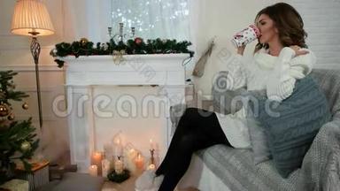 年轻的女人喝茶，快乐的女孩喝茶，微笑着看着镜头，在客厅靠近圣诞树